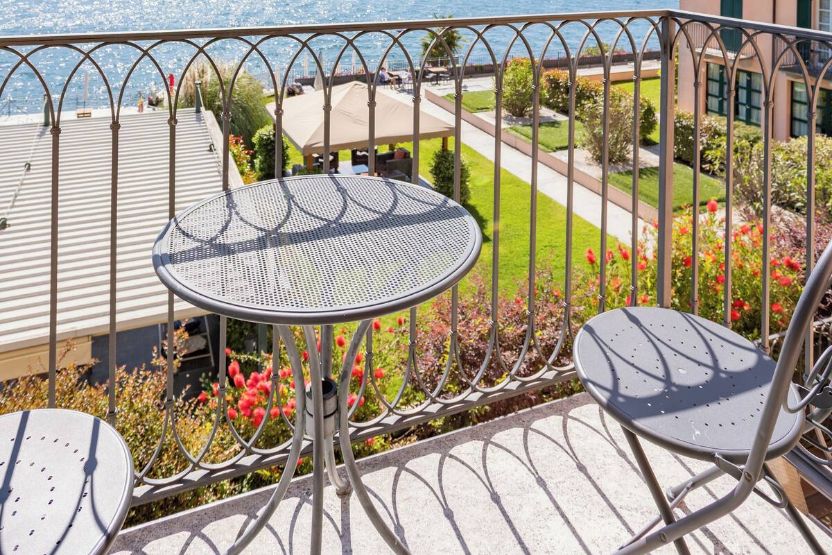 Un balcon avec une table et des chaises en métal, donnant sur une étendue d’eau et une colline lointaine.