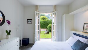 A bedroom with a bed, a dresser, a bedside table with a plant, an open door leading to a garden, and a framed picture on the wall. - Resort Antico Verbano (Meina)