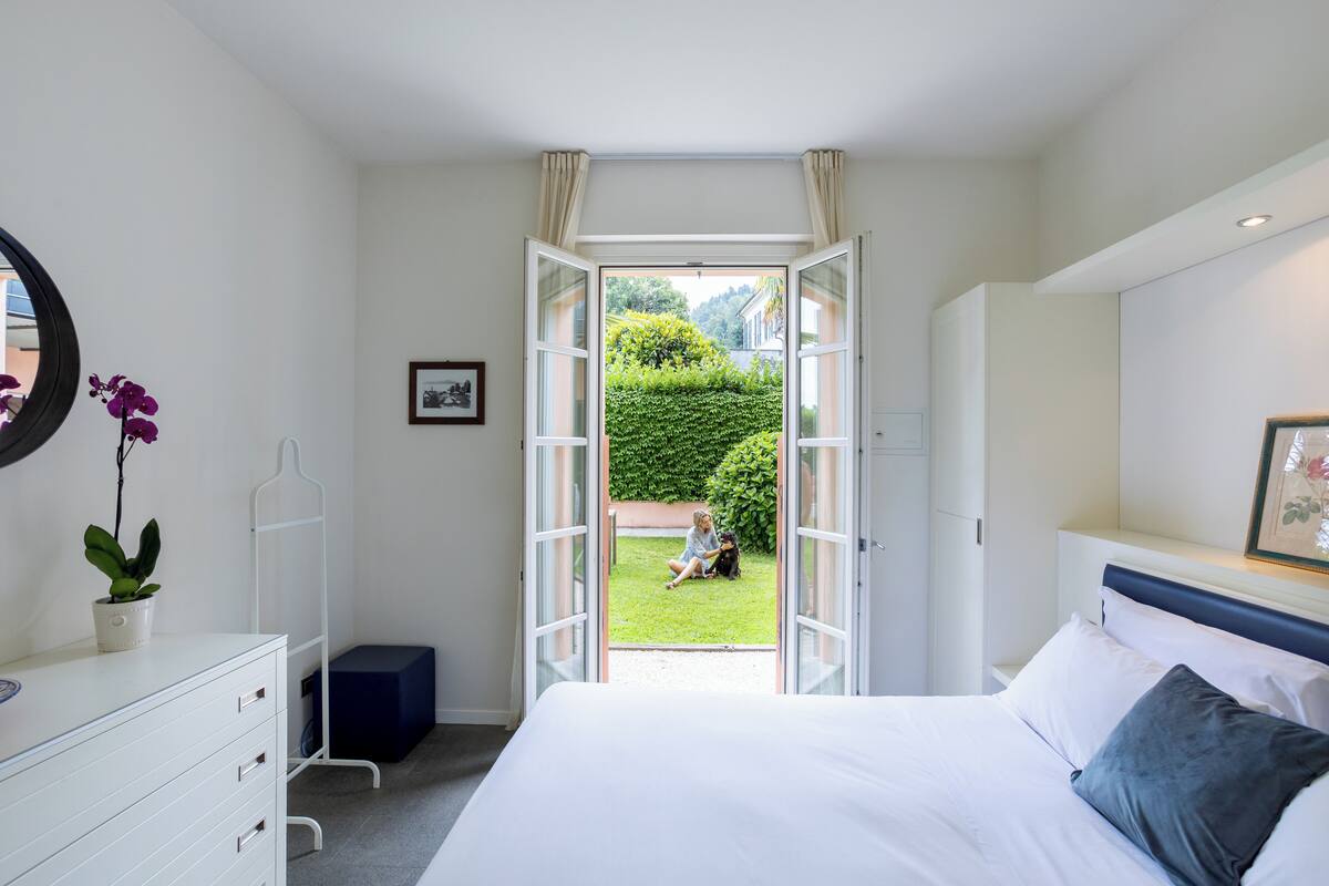 Une chambre à coucher avec un lit, une commode, une table de chevet ornée d’une plante, une porte ouverte donnant sur un jardin et un tableau encadré accroché au mur.