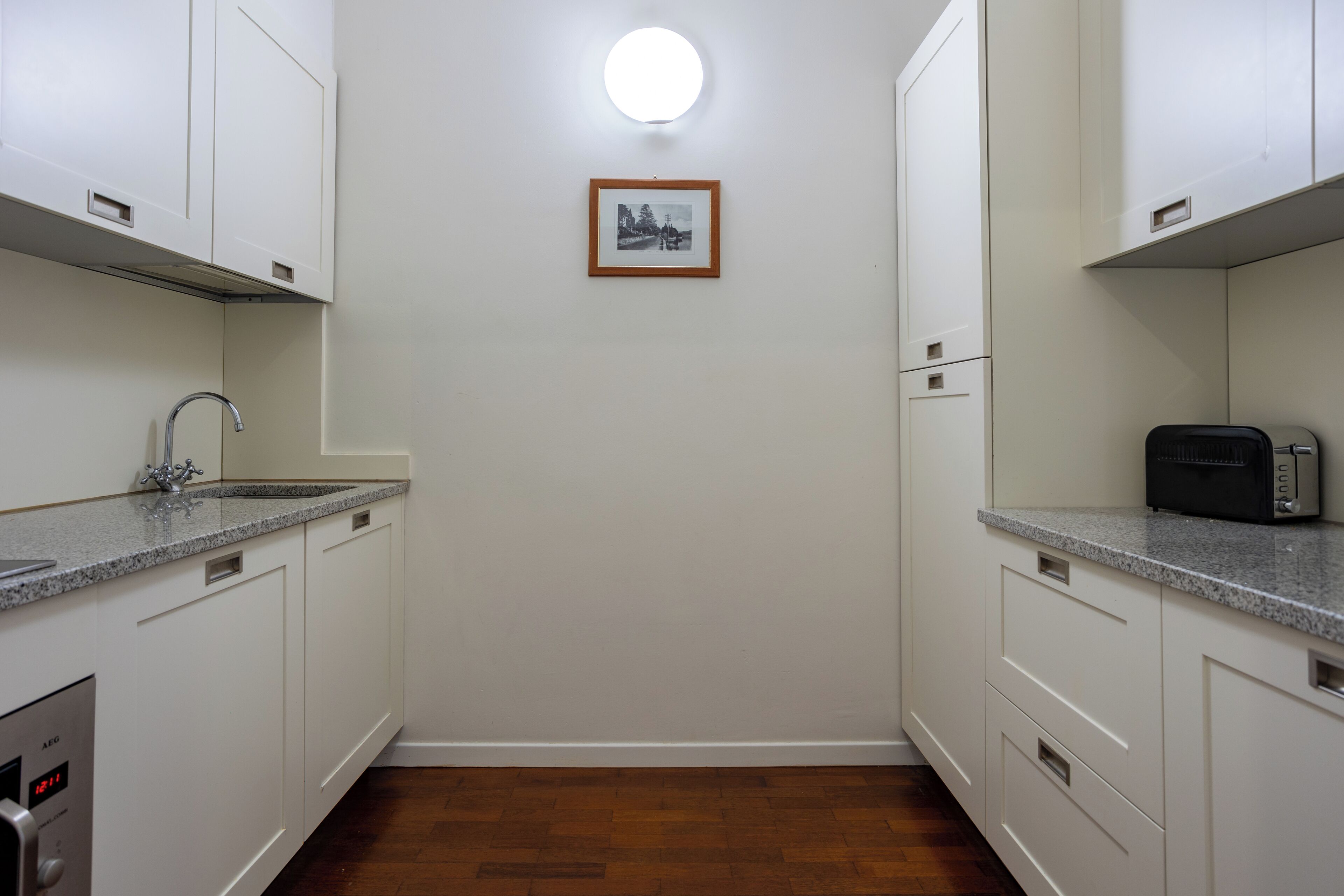 Una cucina compatta con mobili bianchi, un piano di lavoro in granito, forno integrato e una singola foto incorniciata appesa al muro.