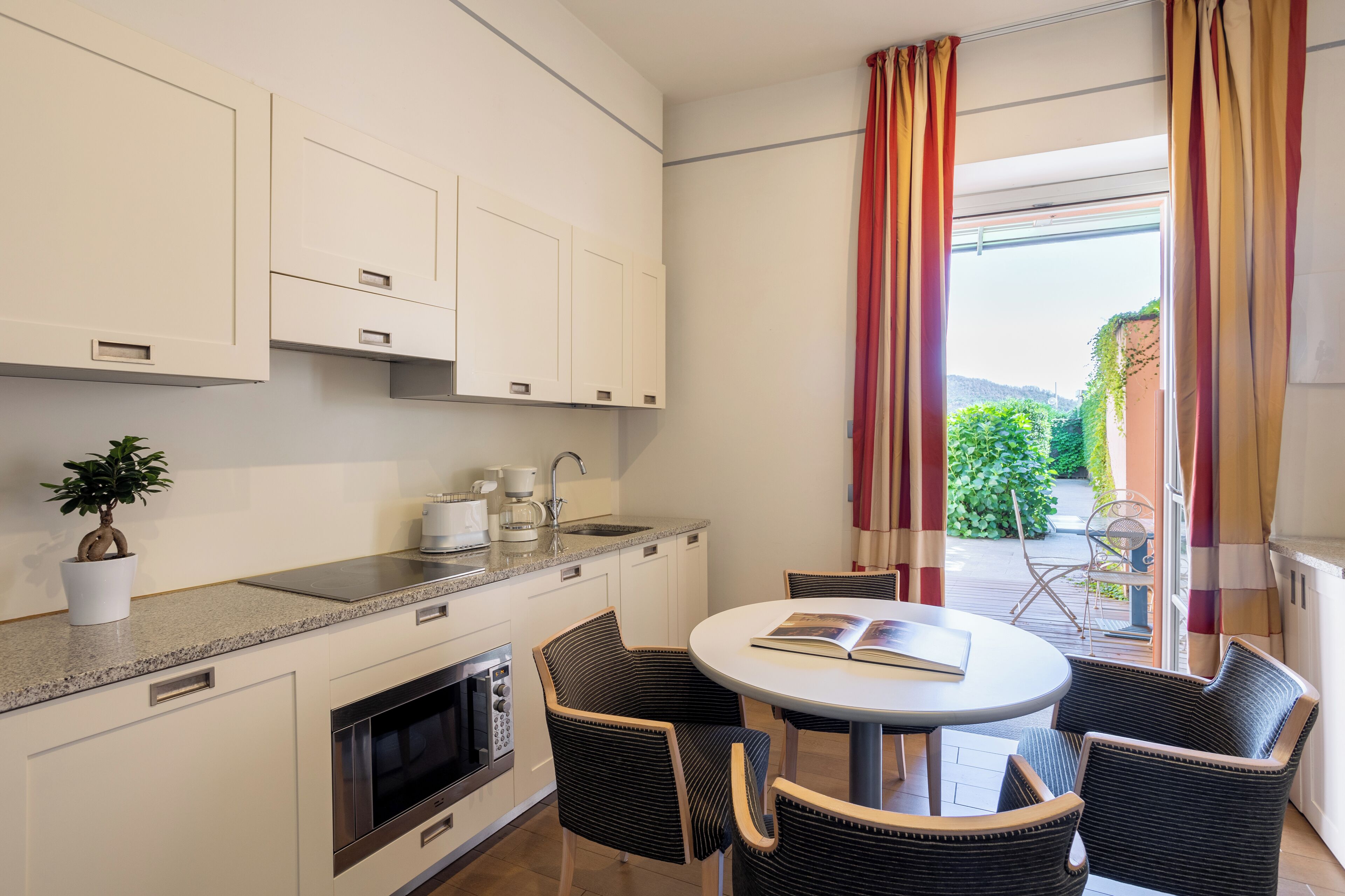 A modern kitchen with white cabinets, a built-in oven, and a coffee maker. There is a round table with four chairs, a potted plant, and a view of a garden through a window with red curtains.