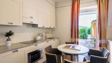 A modern kitchen with white cabinets, a built-in oven, and a coffee maker. There is a round table with four chairs, a potted plant, and a view of a garden through a window with red curtains.