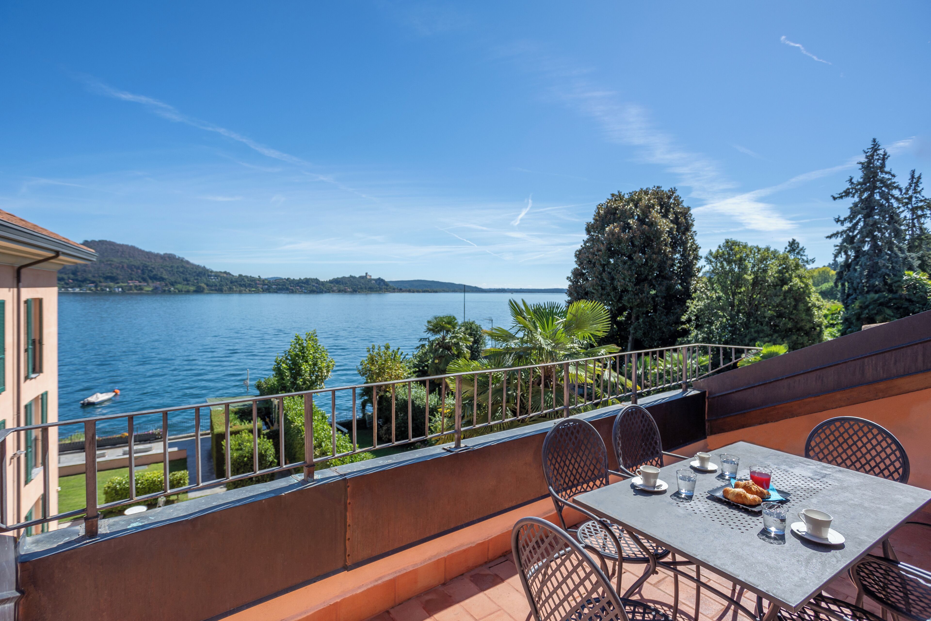 A balcony with a table set for two, overlooking a lake with boats and hills in the distance.