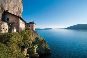 A stone building with arched windows and a tower, situated on a cliff overlooking a calm lake with distant mountains. - Resort Antico Verbano (Meina)
