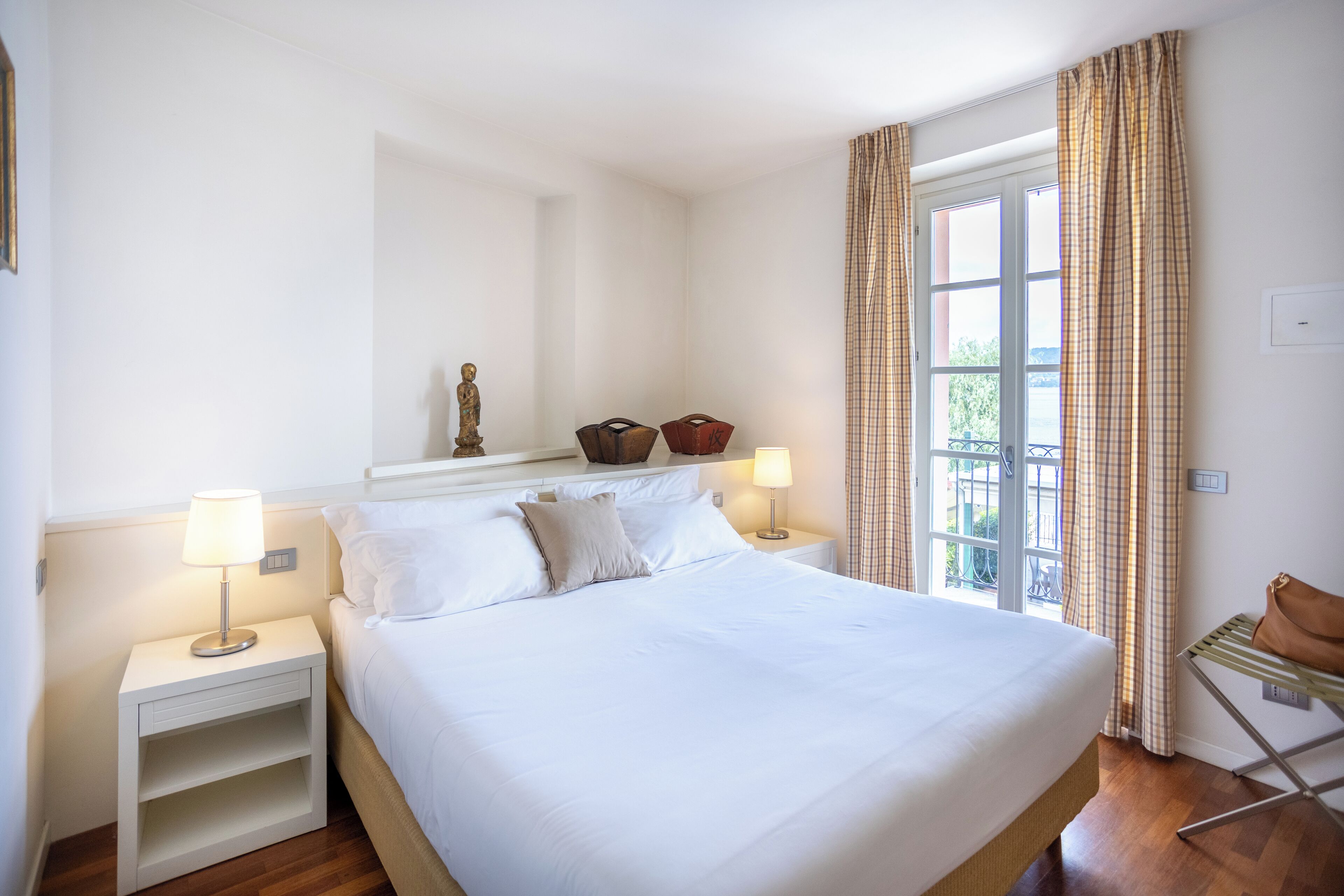 Une chambre bien rangée, avec un grand lit, deux tables de chevet, une commode, une fenêtre avec des rideaux et une petite chaise.