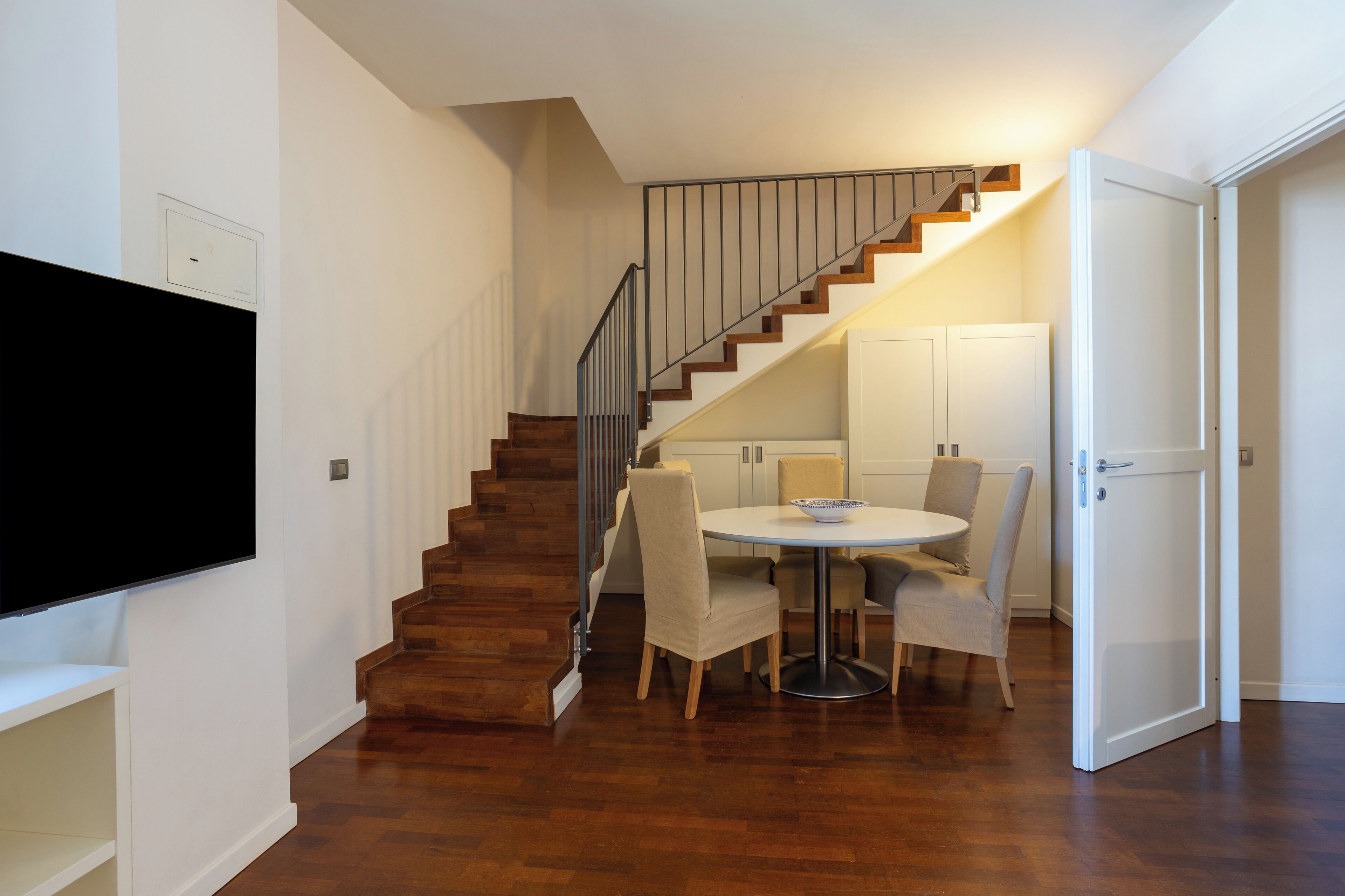 A modern interior with a wooden staircase, a dining area with a round table and chairs, and a flat-screen TV mounted on the wall.