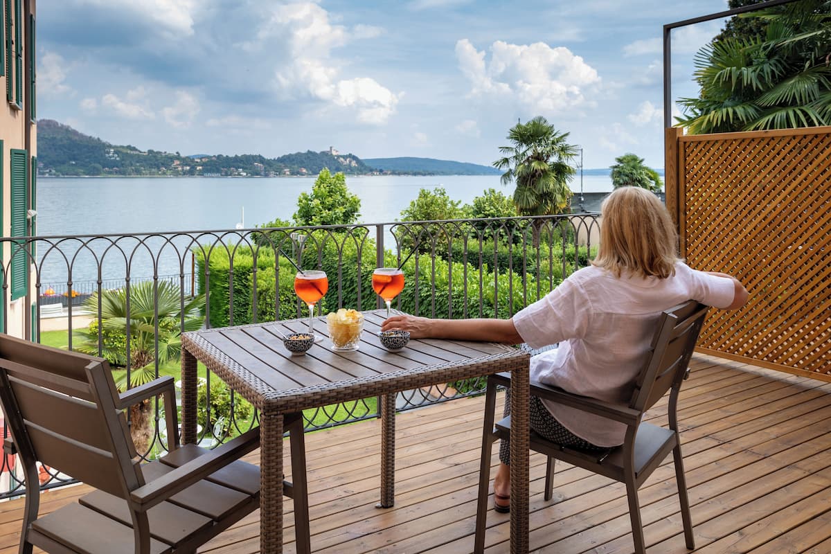 Une personne est assise à une table sur une terrasse en bois donnant sur un lac, avec vue sur des collines et un bâtiment situé au loin.