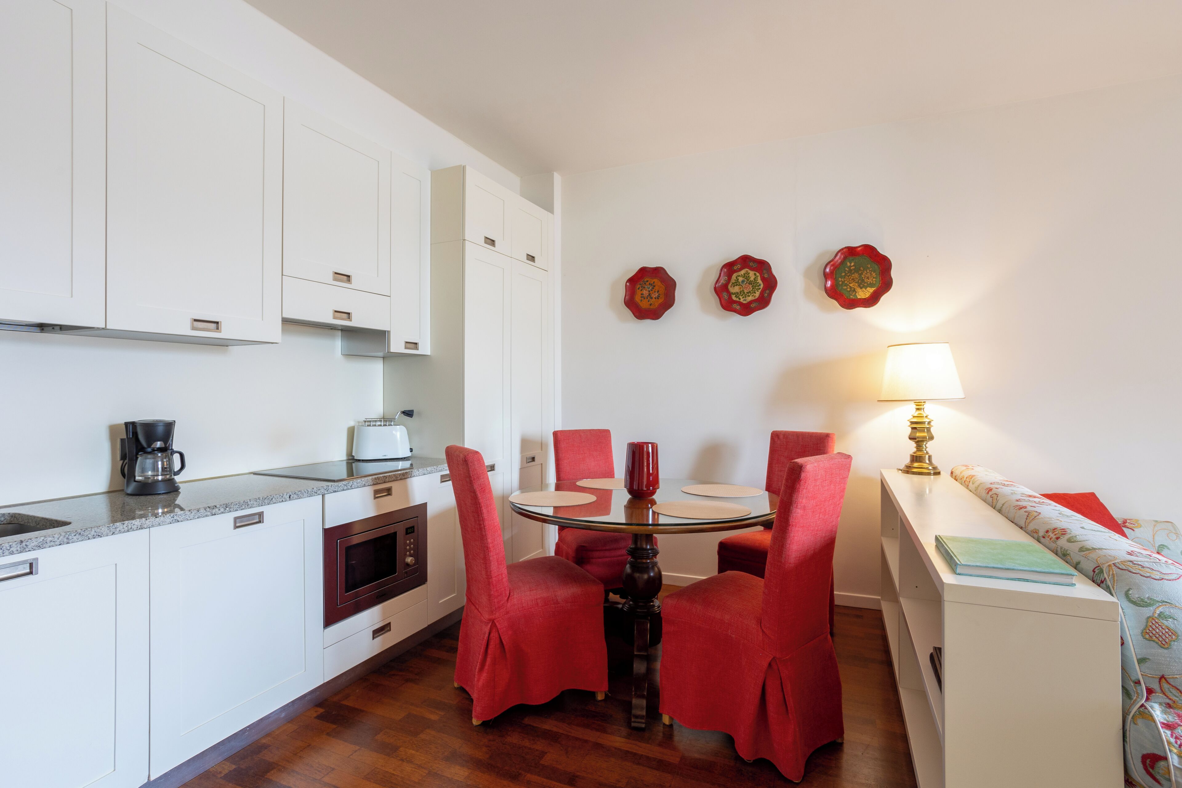 Una cocina compacta con armarios blancos, una cafetera y un tostador. Hay una mesa de comedor con sillas rojas y una lámpara sobre una mesita de noche.