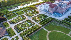 An aerial view of a formal garden with geometric patterns, a large building with a red roof, and a reflective water body. - Resort Antico Verbano (Meina)