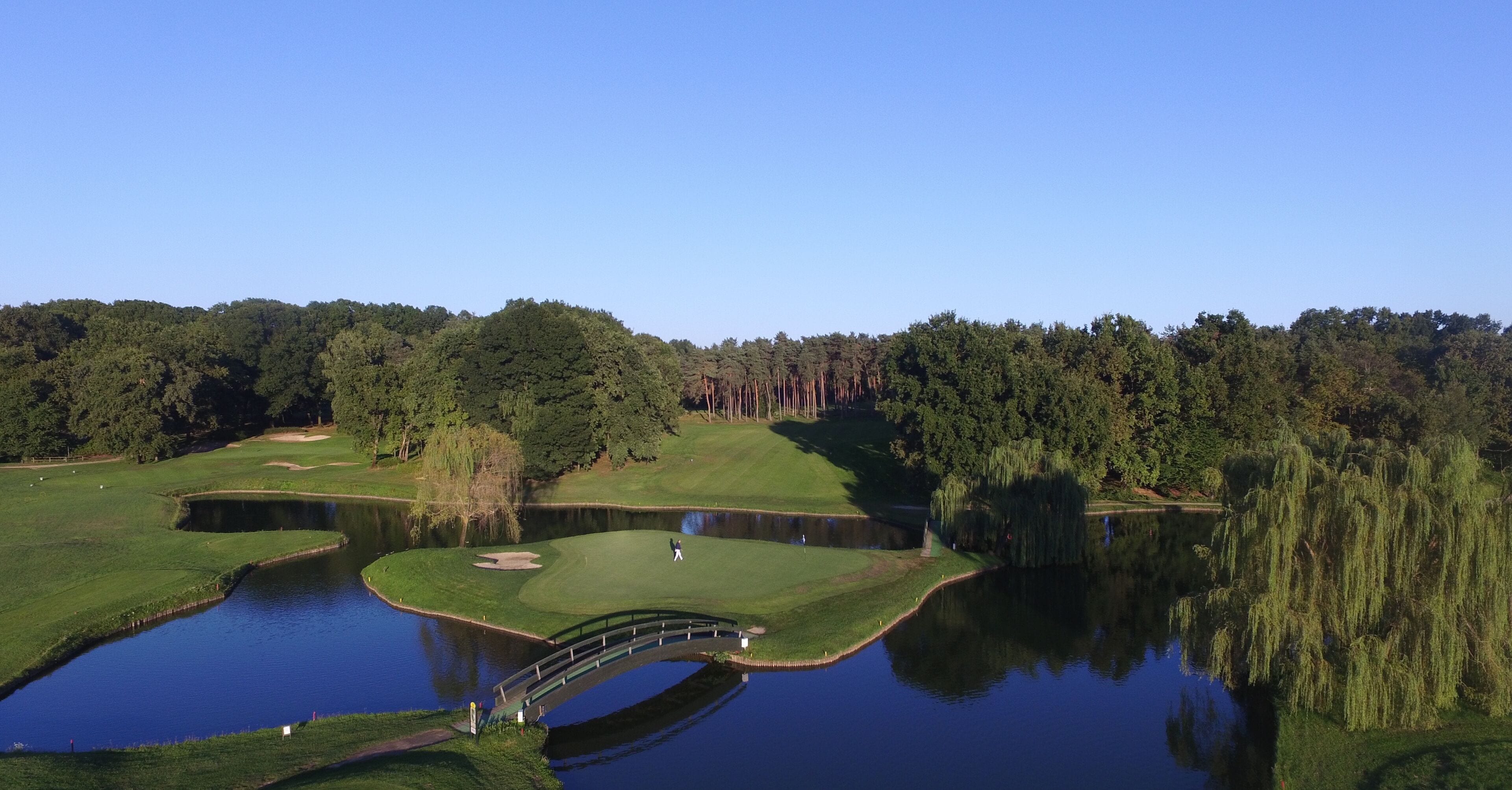 A golf course with a pond, a bridge, and dense forest in the background.