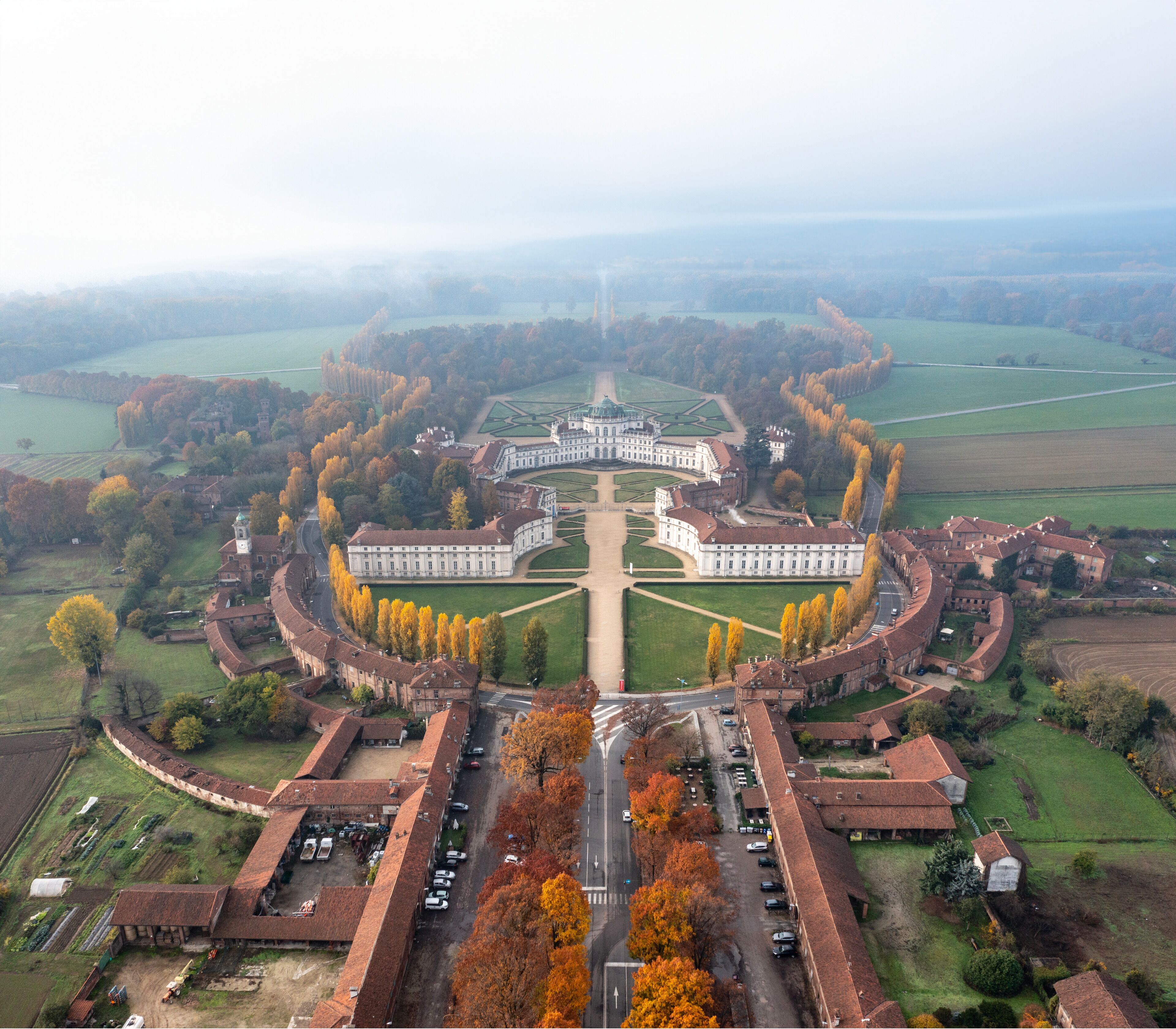 An aerial view of a large, symmetrical building complex with a central courtyard, surrounded by open fields and trees.