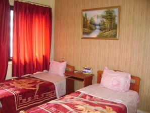 Une chambre d’hôtel avec deux lits, des rideaux rouges, un tableau représentant une forêt et une petite table sur laquelle se trouve une boîte de mouchoirs.