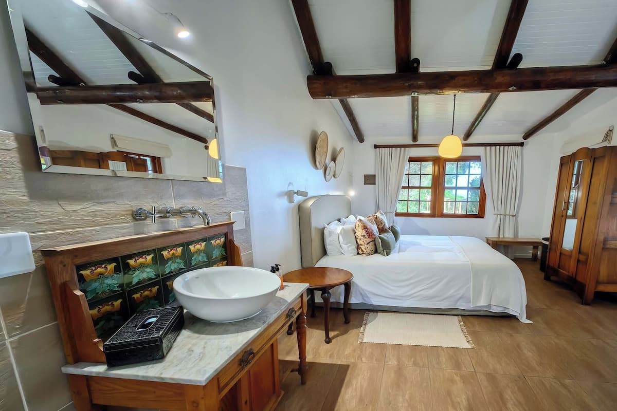 A bedroom with a large bed, wooden beams, a window with curtains, a bedside table, and a bathroom with a sink and mirror.