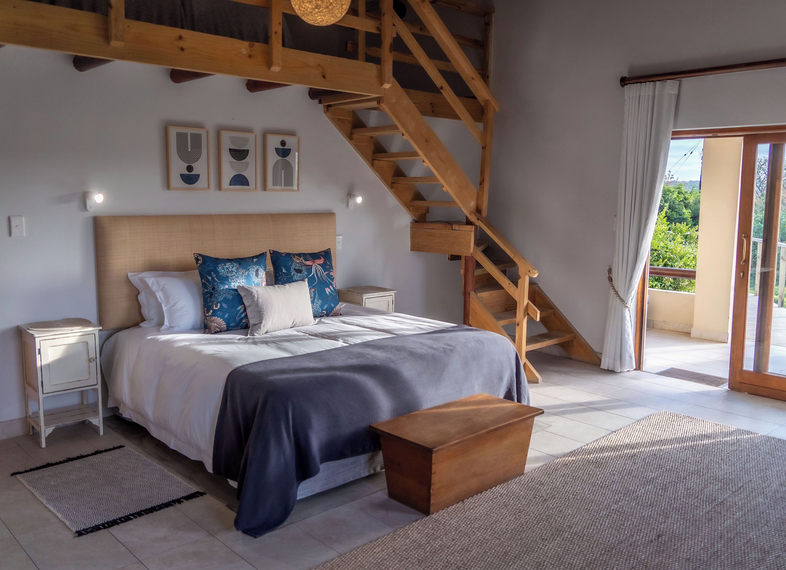 Un dormitorio con una escalera de madera, una cama con almohadones estampados, una mesita de noche y vistas al verde a través de la ventana.