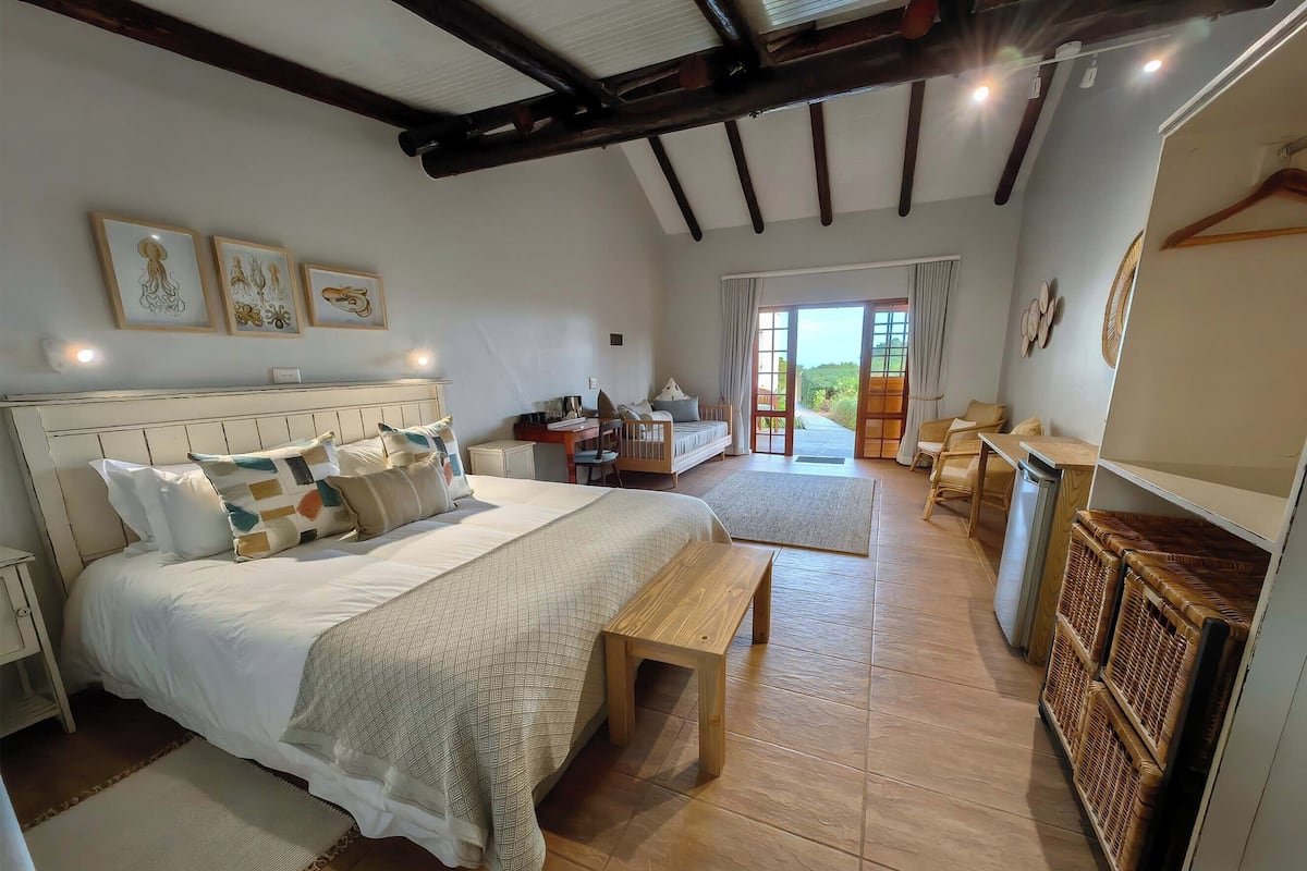 A spacious bedroom with a large bed, a wooden bench, a desk, and a chair. There is a view of the outdoors through a glass door.