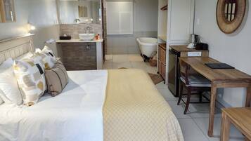 Quarto de hotel com cama, mesa de cabeceira, escrivaninha e banheiro com banheira e pia.
