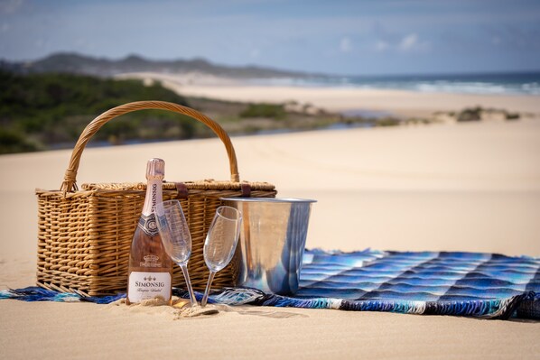 Une scène de plage avec un panier en osier, une bouteille de champagne, deux verres et un seau en métal, le tout posé sur une couverture à motifs bleus et blancs.