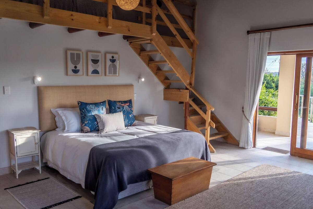 A bedroom with a wooden staircase, a bed with patterned pillows, a bedside table, and a view of greenery through the window.