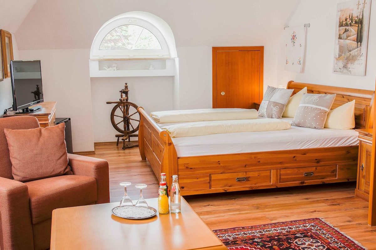 A bedroom with a wooden bed, a desk with a computer, a chair, a small table with glasses and bottles, a window, and a door.