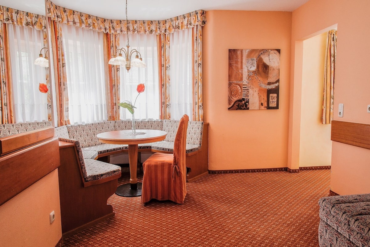 A cozy room with a round table, a sofa, and a chair. There is a painting on the wall and a window with curtains.