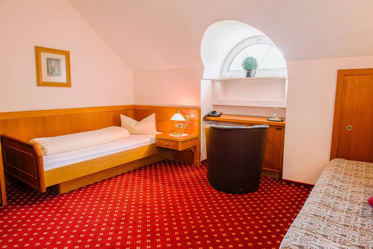 A small hotel room with a single bed, a nightstand with a lamp, a trash bin, and a small kitchenette.