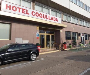 A hotel named 'HOTEL COGULLADA' with a car parked in front, a glass door entrance, and a small outdoor seating area.