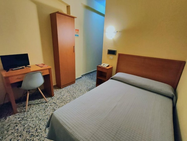 A hotel room with a bed, a desk with a computer, a chair, a wardrobe, and a bedside table with a phone.