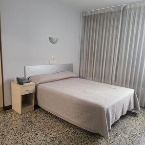A hotel room with a bed, a bedside table with a phone, a wall-mounted light, and a window with curtains.