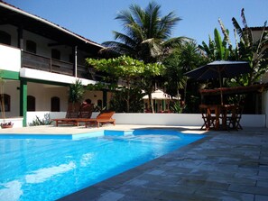 Une piscine entourée de palmiers et d’un bâtiment doté d’un balcon, avec des chaises longues et des parasols à proximité.