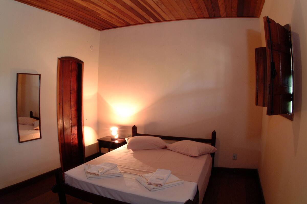 A bedroom with a wooden ceiling, a bed with white linens, a nightstand with a lamp, and a mirror on the wall.