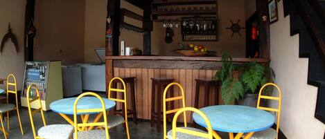 A bar area with wooden tables and yellow chairs, a fruit bowl on the counter, and a potted plant.