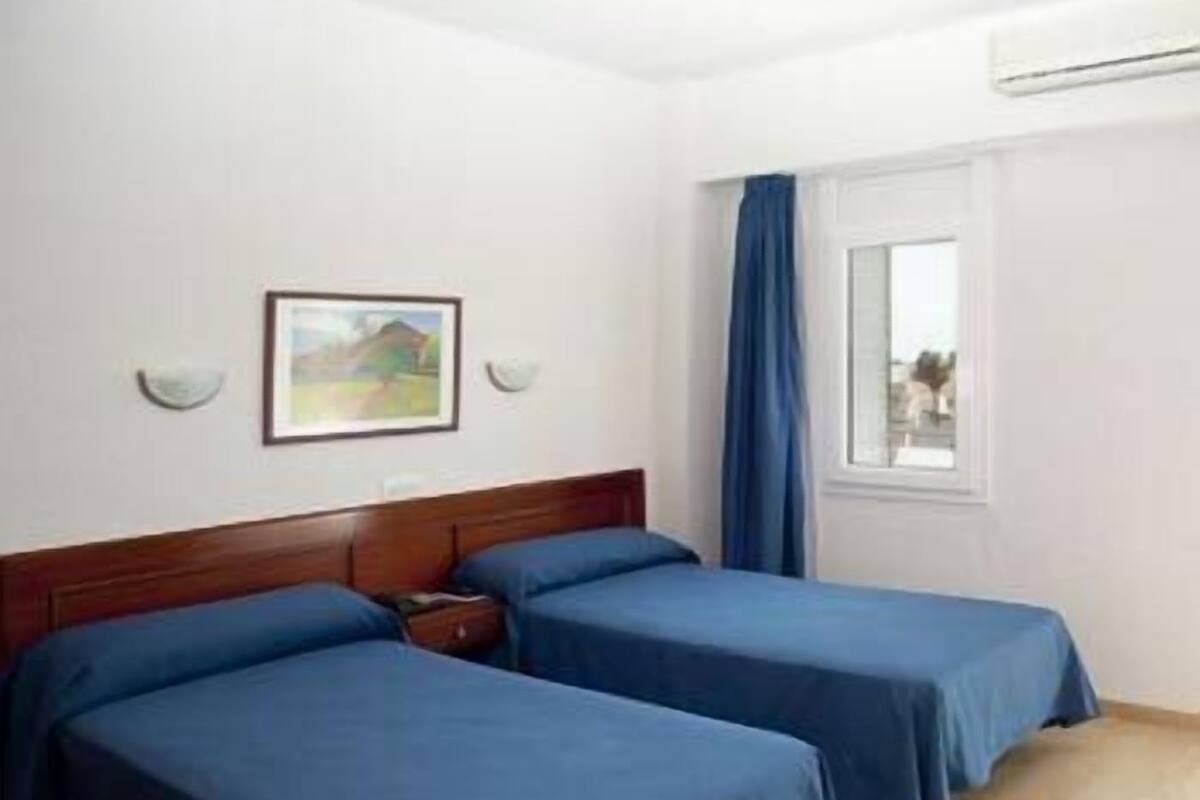 A hotel room with two single beds, a wooden headboard, a window with blue curtains, and a painting on the wall.