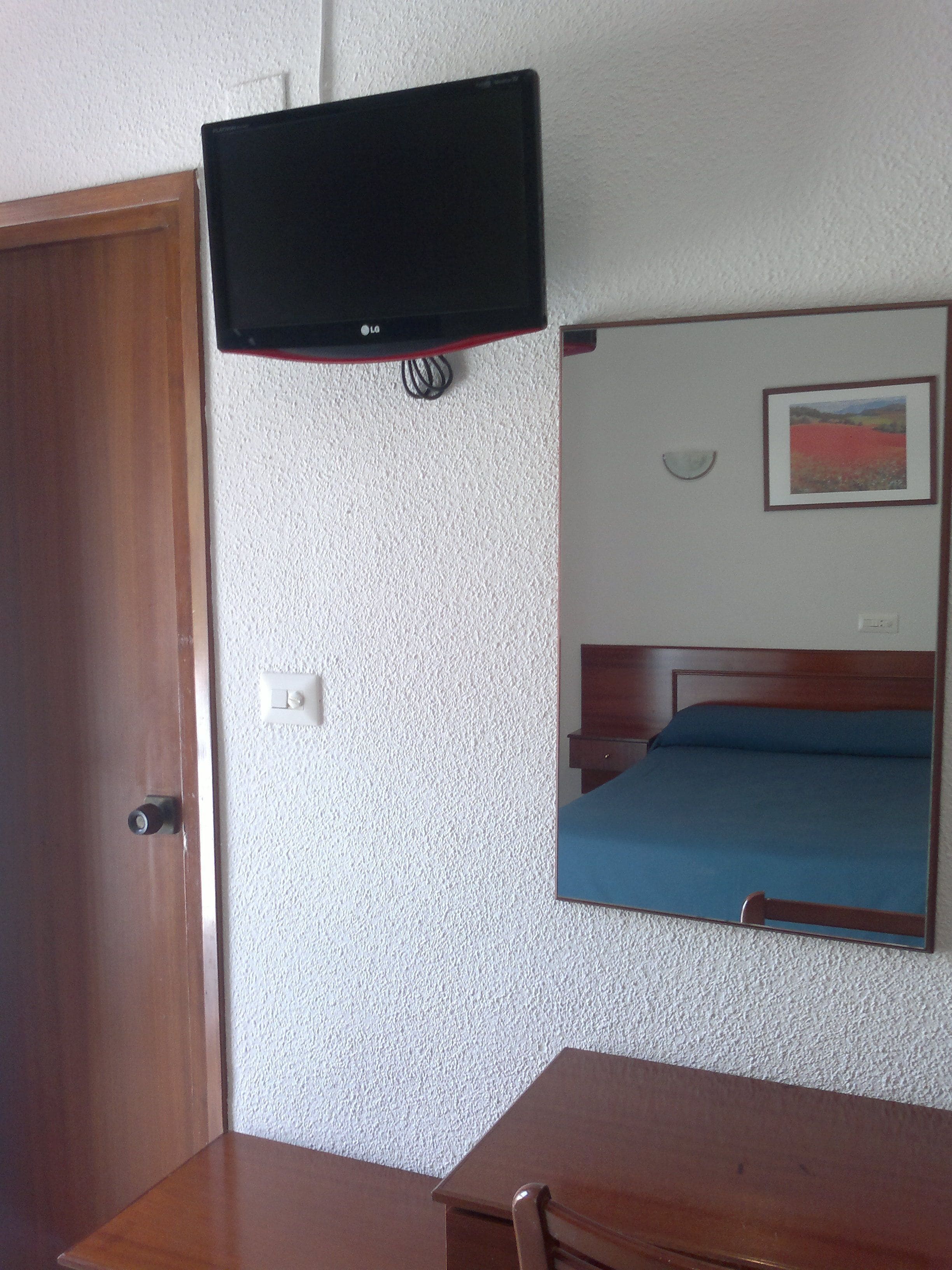 A room with a wooden door, a television mounted on the wall, a bed with a blue mattress, and a framed picture on the wall.