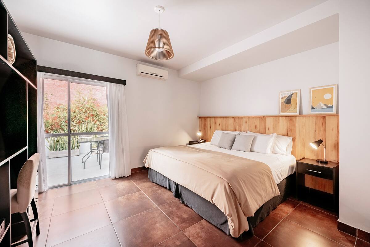 A modern bedroom with a large bed, a wooden headboard, a nightstand with a lamp, and a sliding glass door leading to an outdoor area.