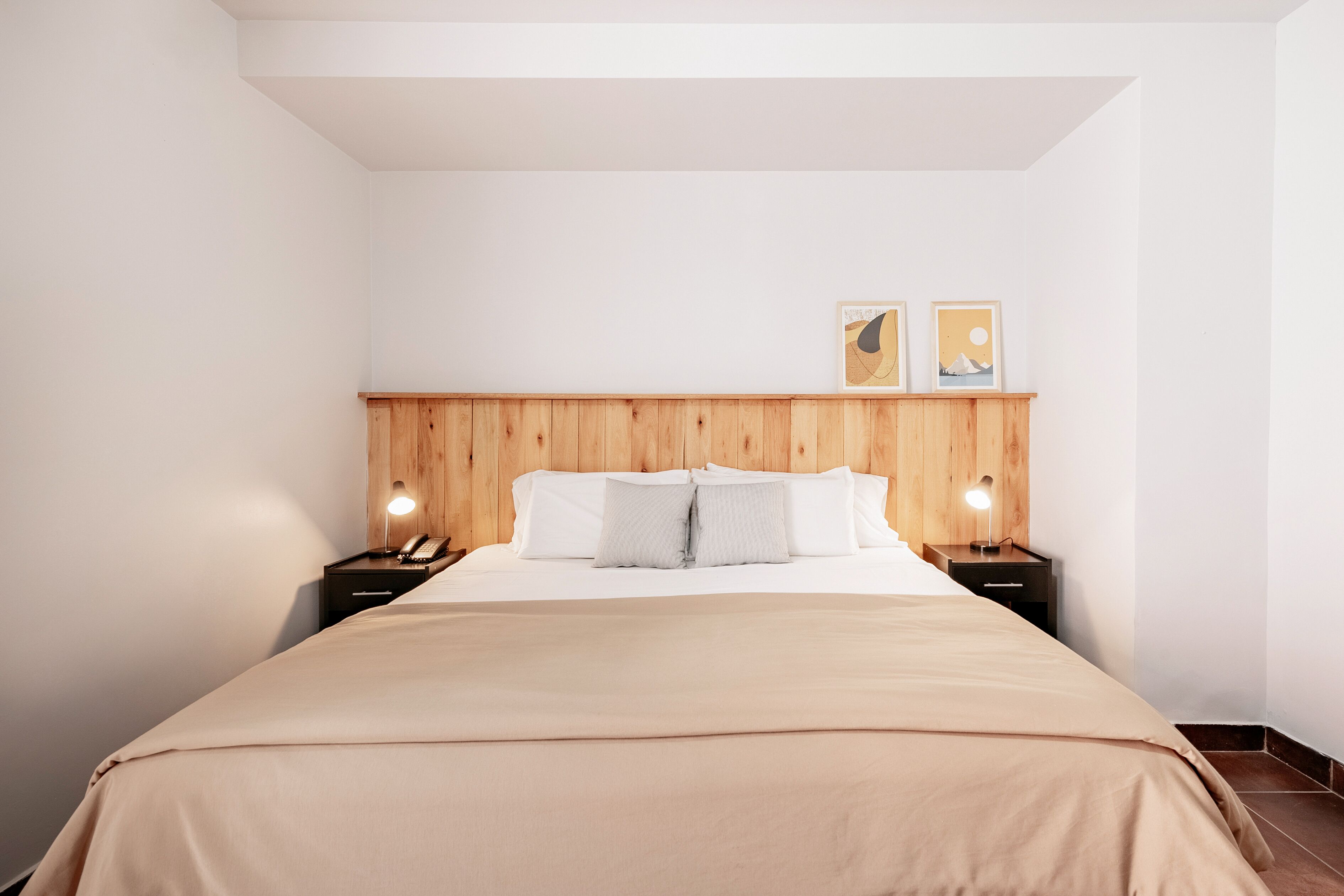 A hotel room with a wooden headboard, a bed with white linens, two bedside tables with lamps, and two framed pictures on the wall.