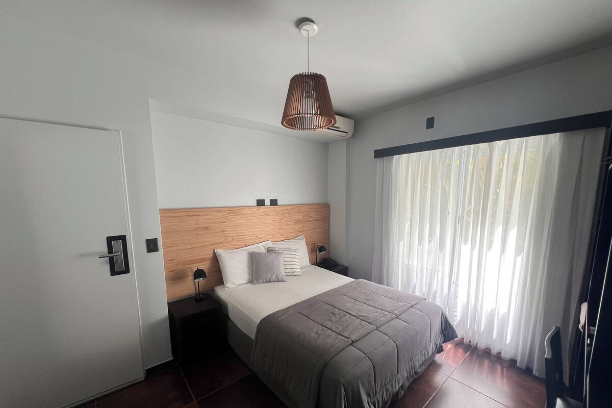 A modern hotel room with a wooden headboard, a large bed with a grey bedspread, a bedside table with a lamp, and a window with sheer curtains.