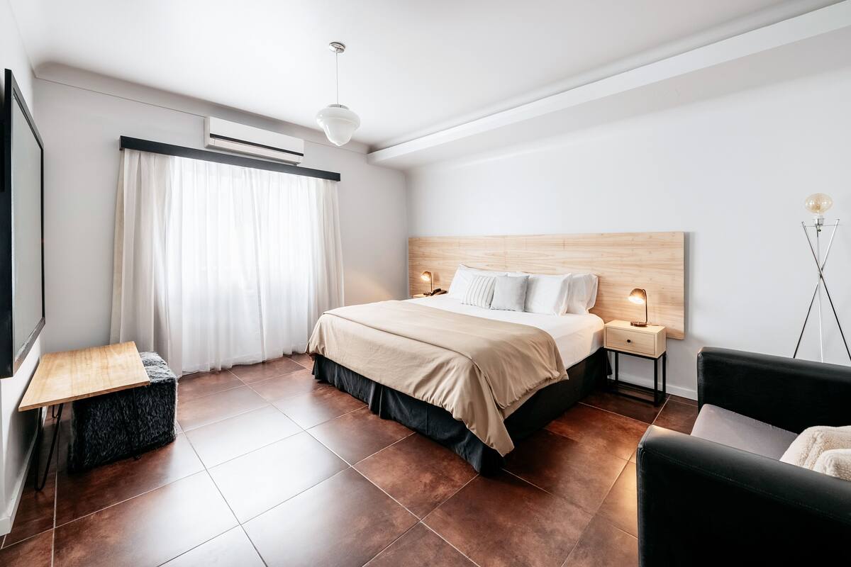 A modern bedroom with a wooden headboard, a bed with a beige cover, a nightstand with a lamp, and a black leather chair.