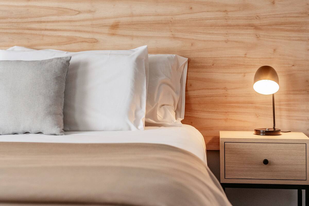A neatly made bed with a wooden headboard, a bedside table with a lamp, and a drawer.