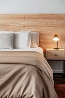 Una cama bien tendida con cabecera de madera, una mesita de noche con lámpara y un cajón.
