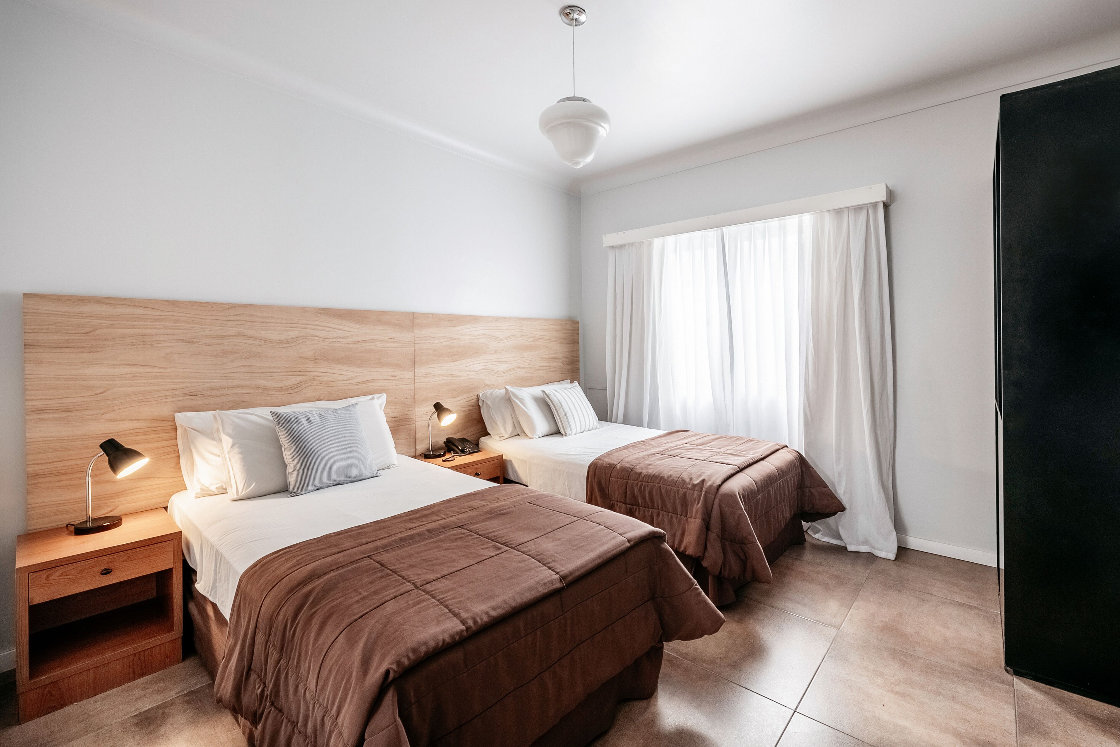 A hotel room with two beds, a wooden headboard, a nightstand with a lamp, and a large window with curtains.