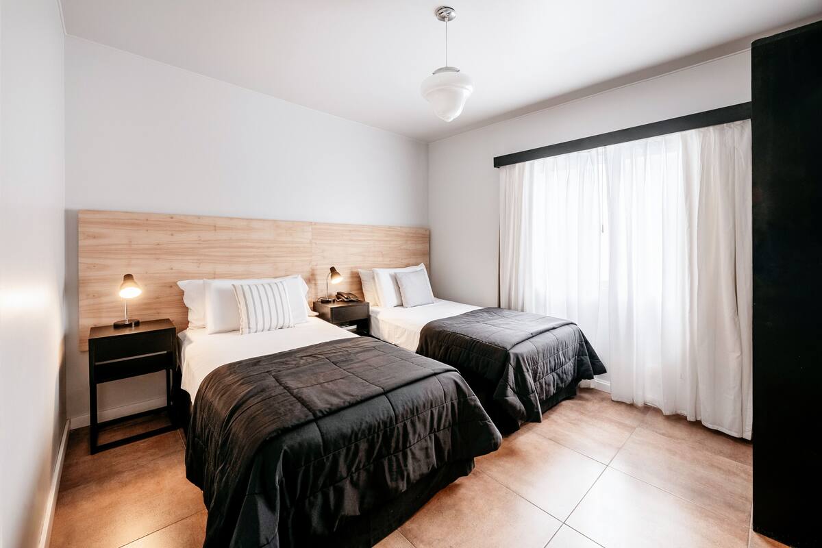 A hotel room with two beds, a wooden headboard, a nightstand with a lamp, and a large window with curtains.