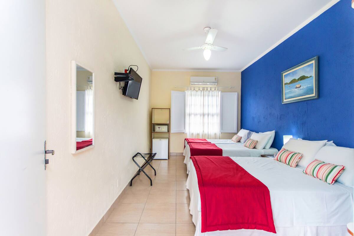 A hotel room with three beds, a TV mounted on the wall, a ceiling fan, and a painting on the blue wall.