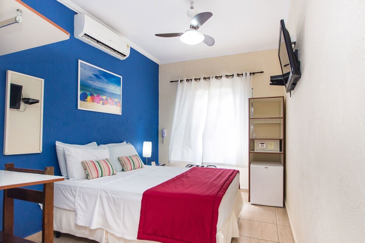 A hotel room with a bed, a desk, a chair, a TV, an air conditioner, a ceiling fan, a window with curtains, and a shelf with items.