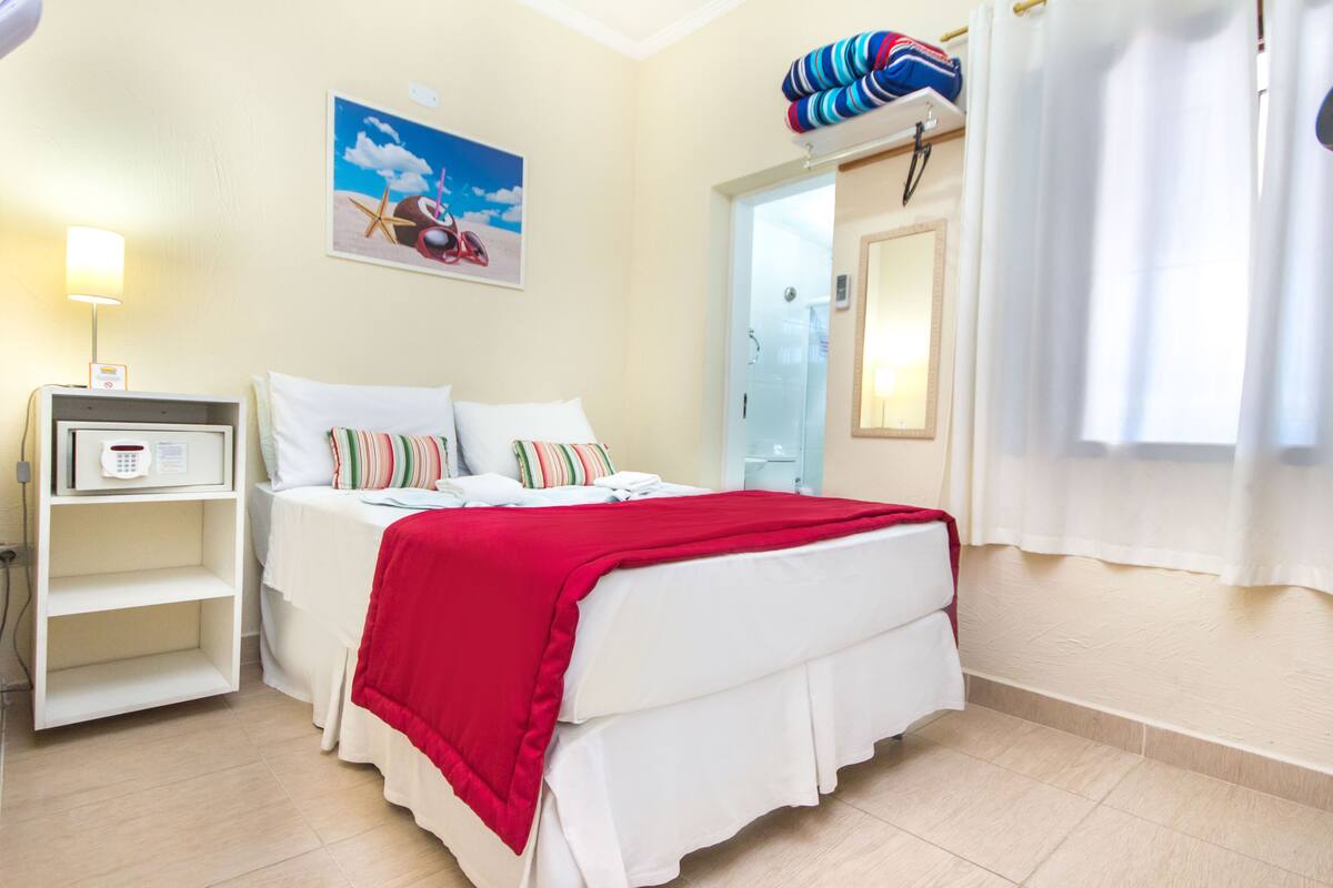 A hotel room with a bed, a bedside table, a lamp, a painting, and a window with curtains.