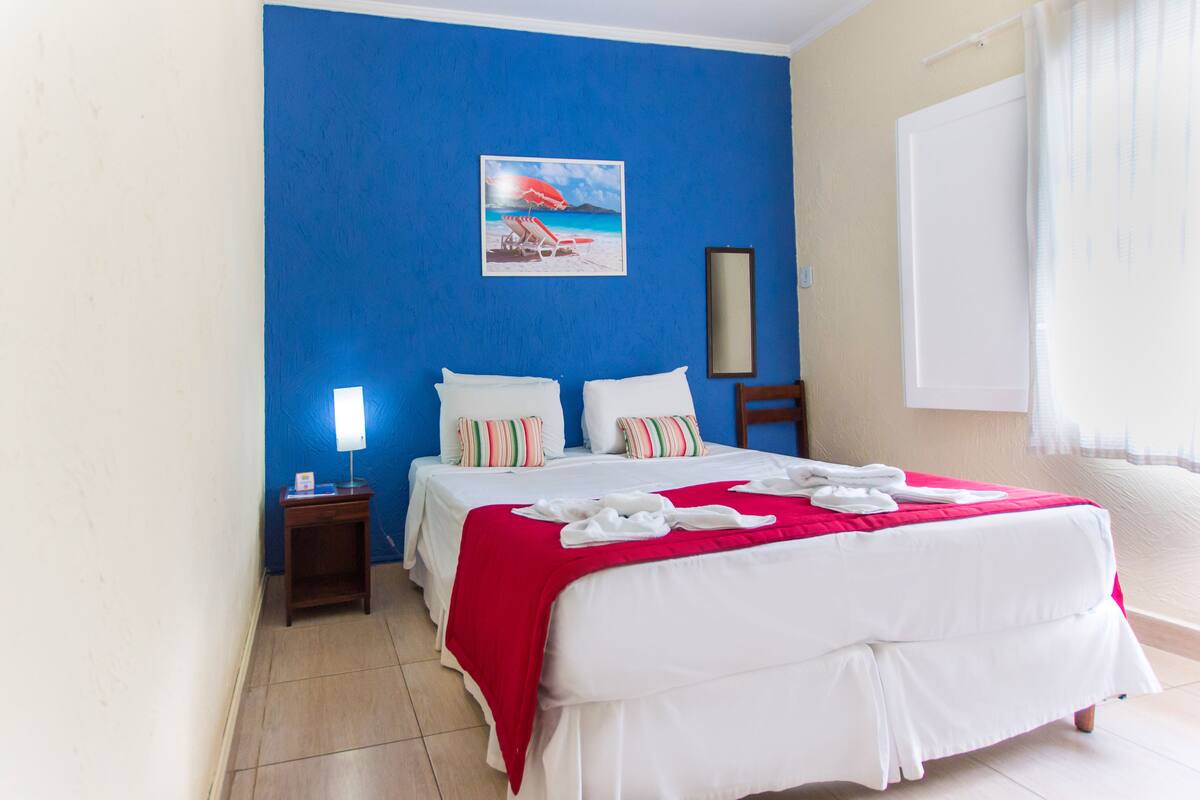 A hotel room with a large bed, a nightstand, a painting of a beach scene, and a window with curtains.