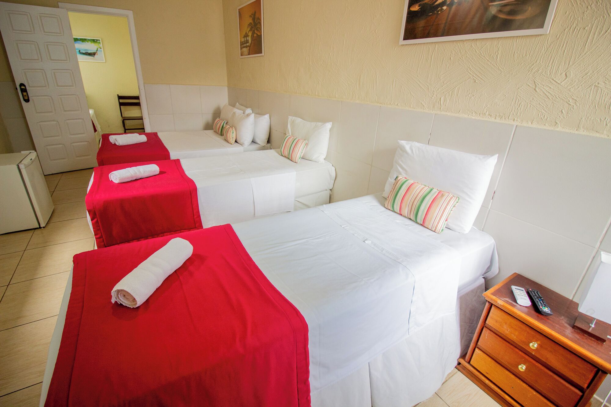 Quarto de hotel com duas camas, cada uma com colcha vermelha e almofadas brancas, mesa de cabeceira de madeira com um controle remoto, e uma porta com vista para o exterior.
