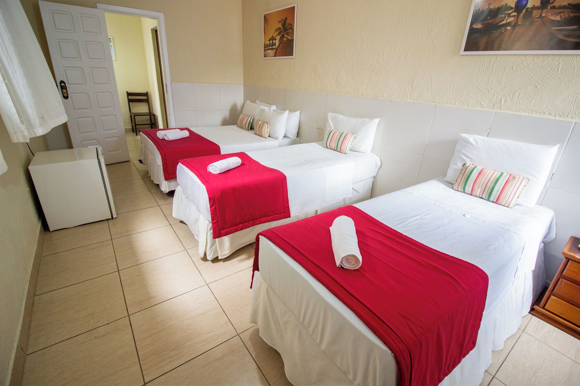 Habitació d'hotel amb quatre llits, cada un amb llençols blancs i manta vermella, un refrigerador petit de color blanc i una nitrera de fusta.