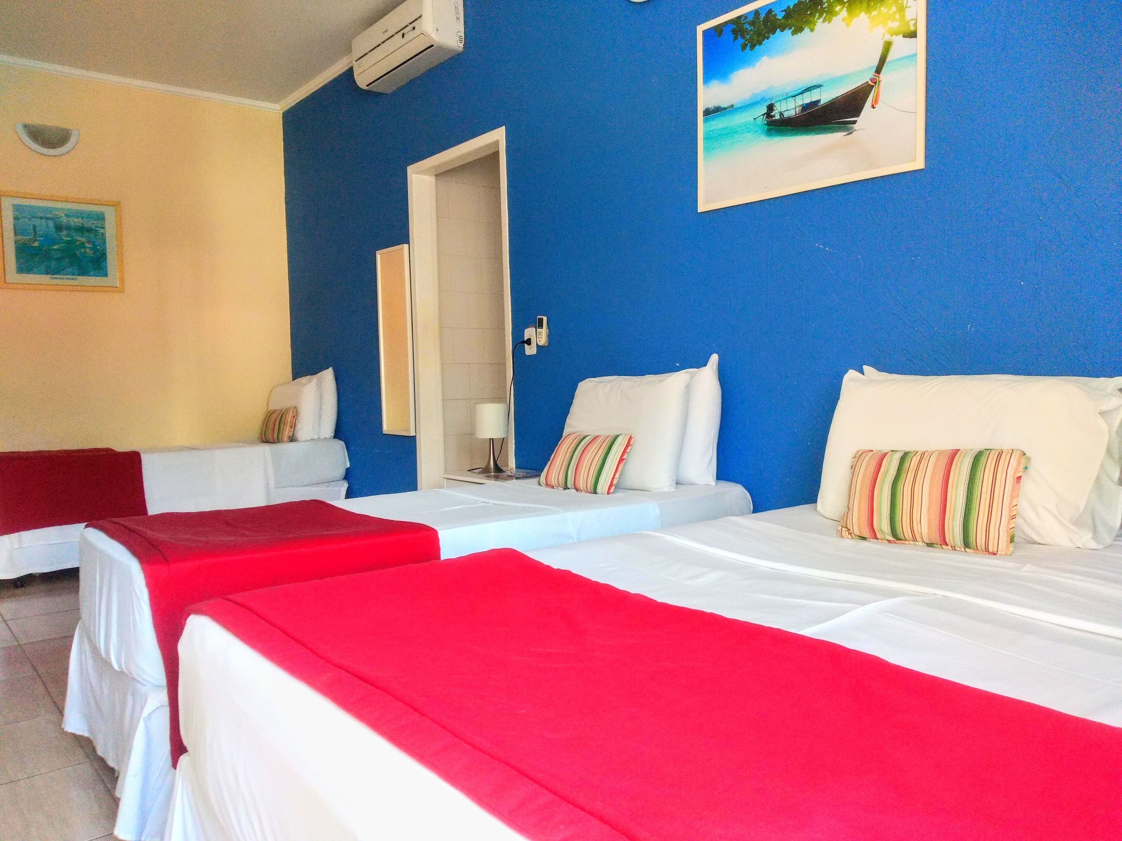 Ein Hotelzimmer mit zwei Betten, jeweils mit roter Bettdecke, weißen und gestreiften Kissen. An der Wand hängt ein Bild eines Bootes, auf dem Nachttisch steht ein Telefon und eine Tür führt in einen anderen Raum.
