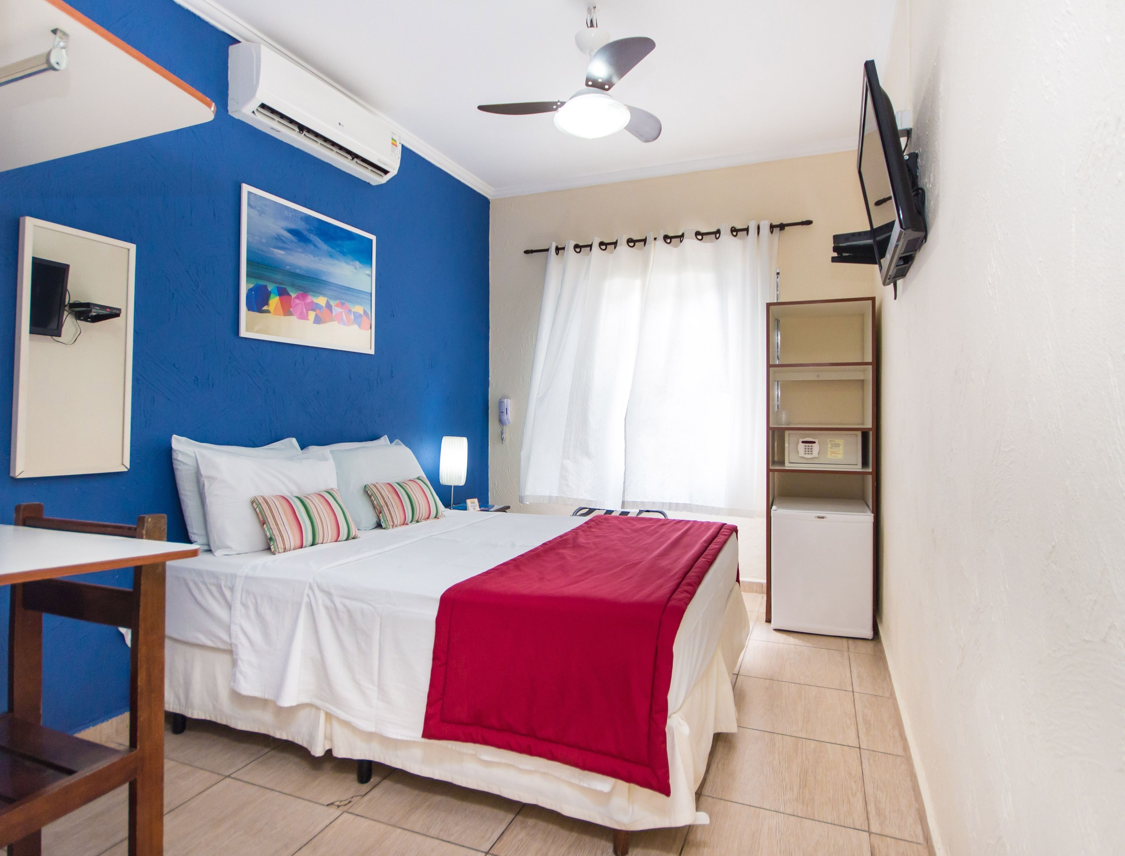 A hotel room with a bed, a desk, a chair, a TV mounted on the wall, and a ceiling fan.