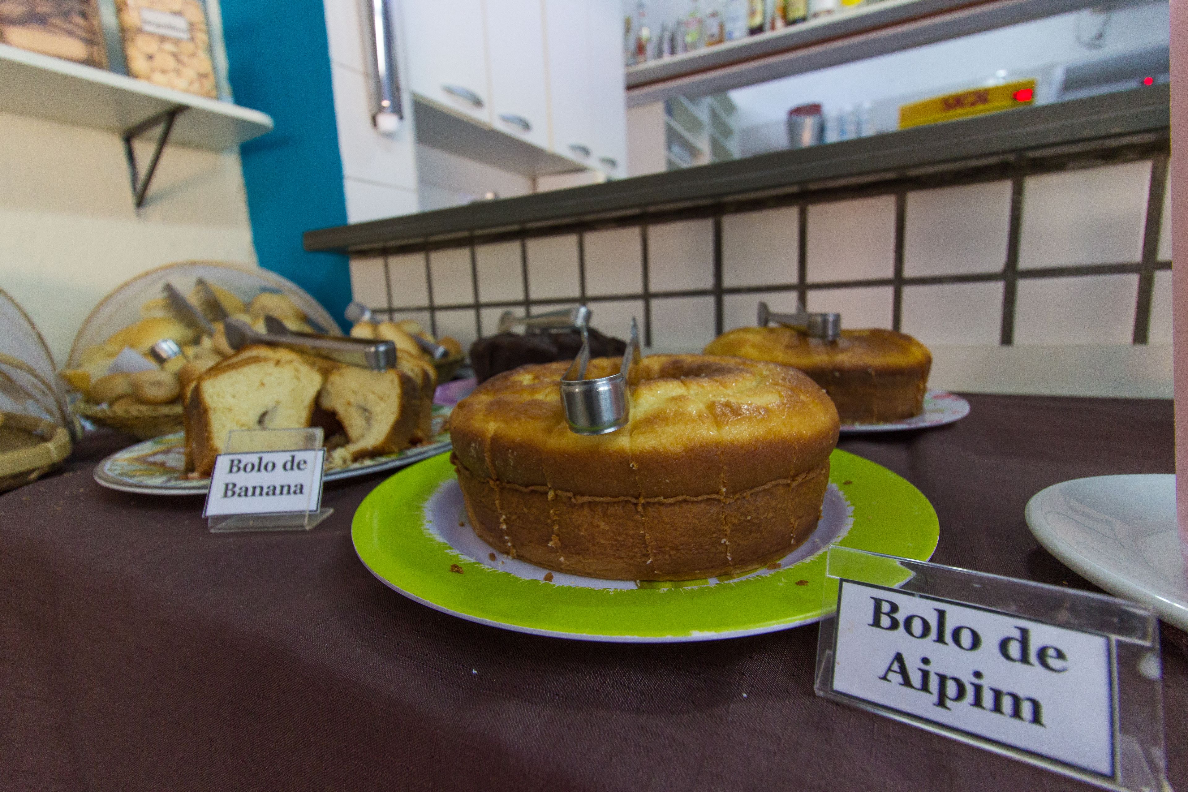 Una exhibición de pasteles con etiquetas, incluyendo “Bolo de Banana” y “Bolo de Aipim”, sobre una mesa en una panadería o cafetería.